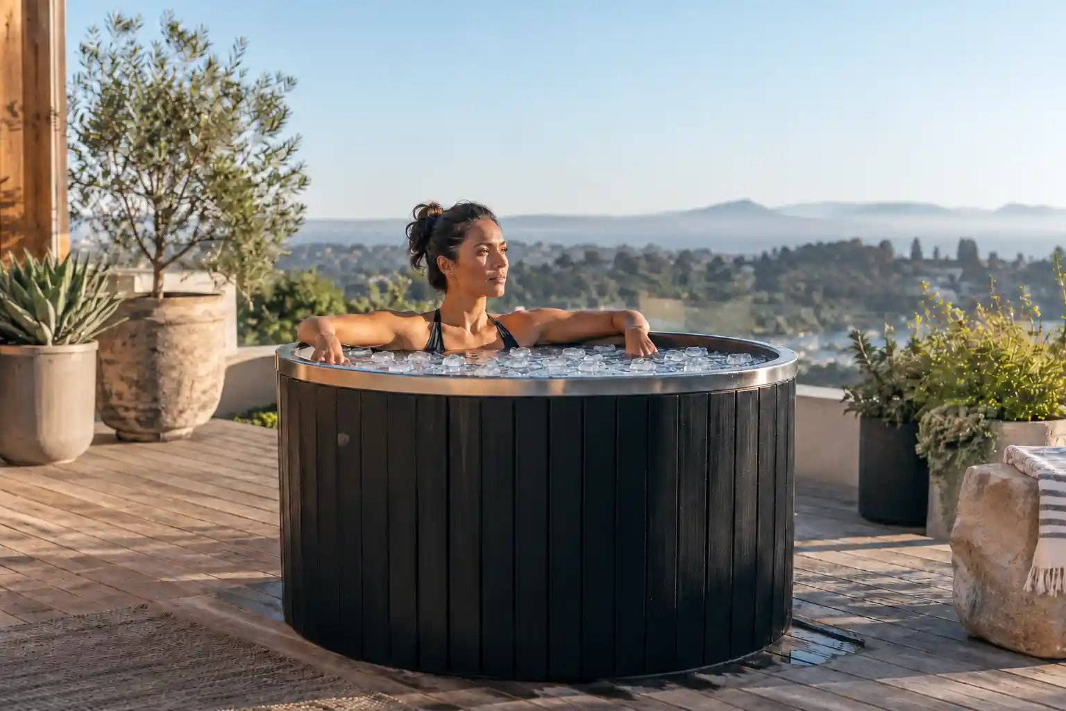 Woman resting her arms on a round black cold plunge barrel with small ice pieces on a sunny hillside terrace.